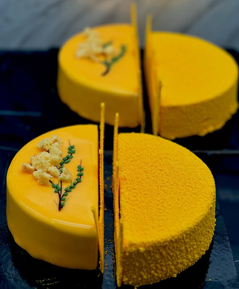 Due torte gialle divise a metà, glassa lucida e superficie vellutata, con decoro a mimosa su ardesia scura